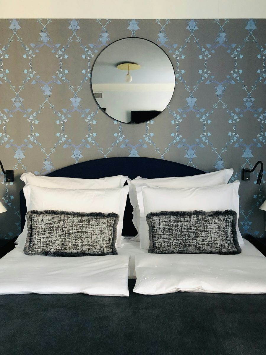 Bedroom with a soft blue damask wallpaper and a round mirror above a dark upholstered headboard